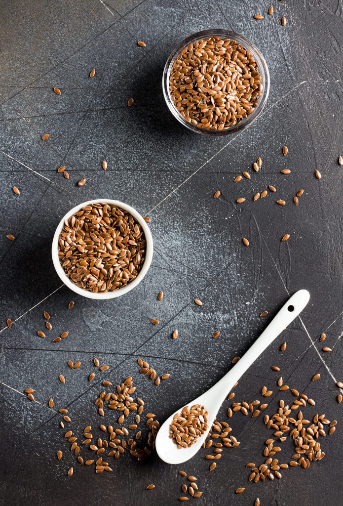 وعاء شفاف من بذور الكتان ووعاء اخر بلون ابيض كذلك يحوي بذور الكتان وملعقة بيضاء فيها بذور وحبوب منثورة على الارضية