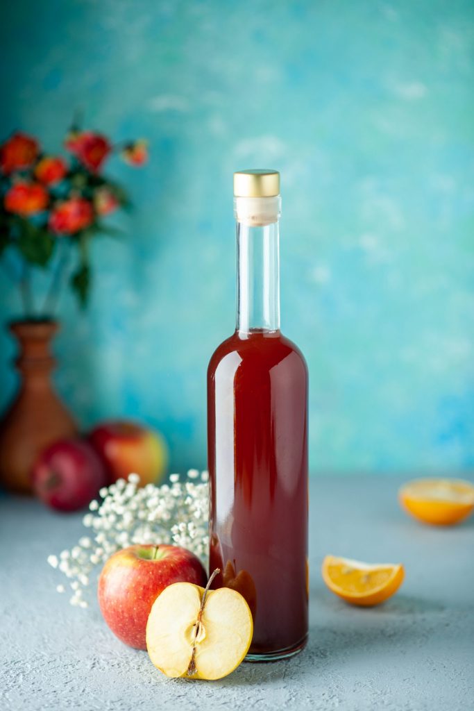 قارورة زجاجية تحتوي على خل التفاح فوق طاولة وبجانبها يوجد تفاح أحمر وعرق ورد أبيض ووراء القارورة بجانب احائط يوجد مزهرية ورد وتفاحتين وقطع برتقال اورانج